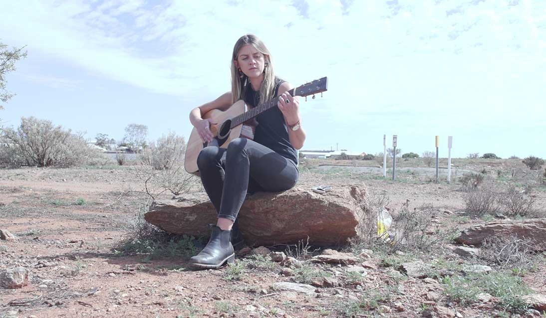 Woman sits on a tree stump playing a guitar.