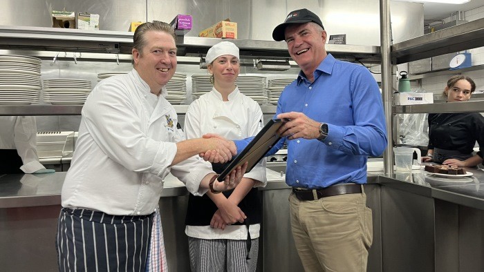 A man presents a certificate to a chef dressed in white clothes and an apron while another person looks on