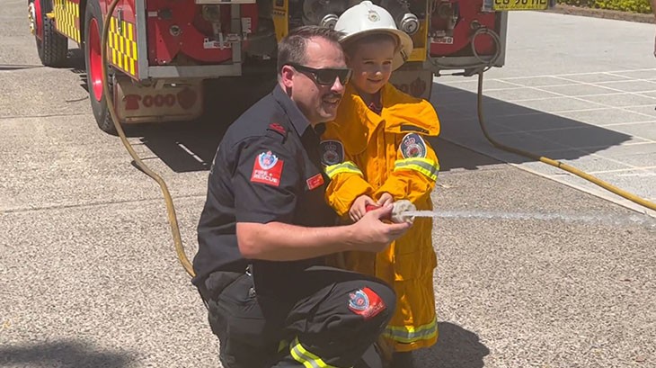 A young boy gets help from a firefighter to point a fire hose.