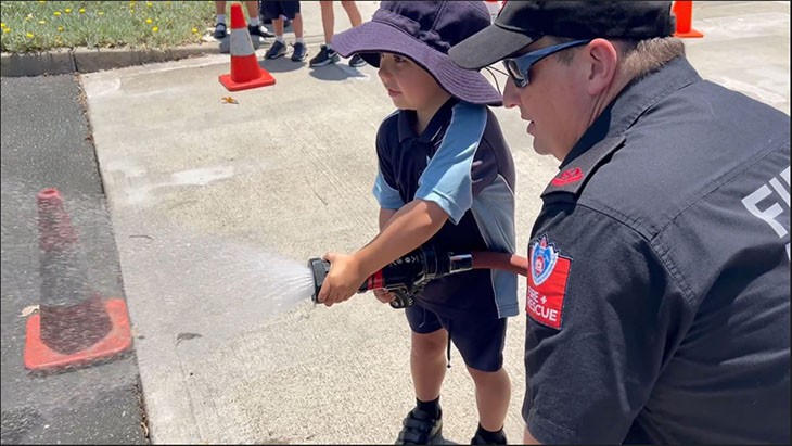 A little boy holds a fire hose while a firefighter watches on.