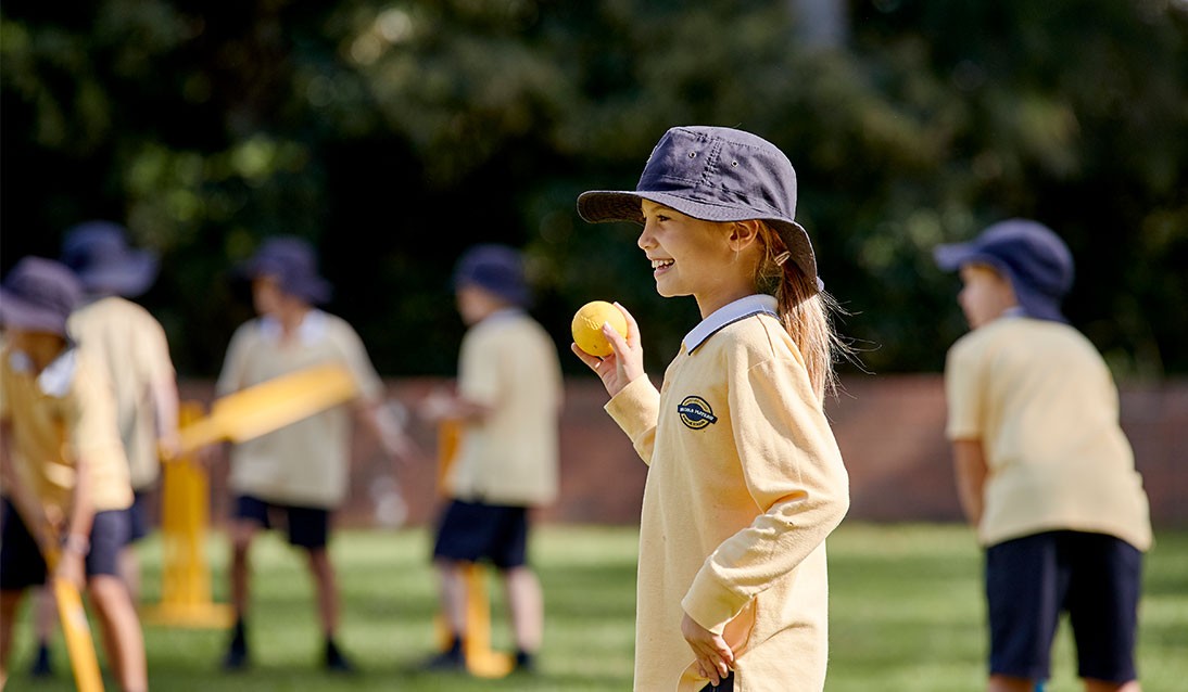 A primary school girl bowls a cricket ball while other students play with bats in the background.