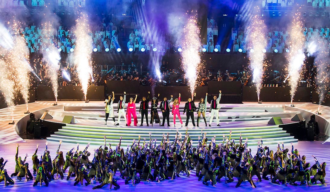 Student performers stand on stage with coloured lights and a pyrotechnic display behind them in an image from the 2019 Schools Spectacular