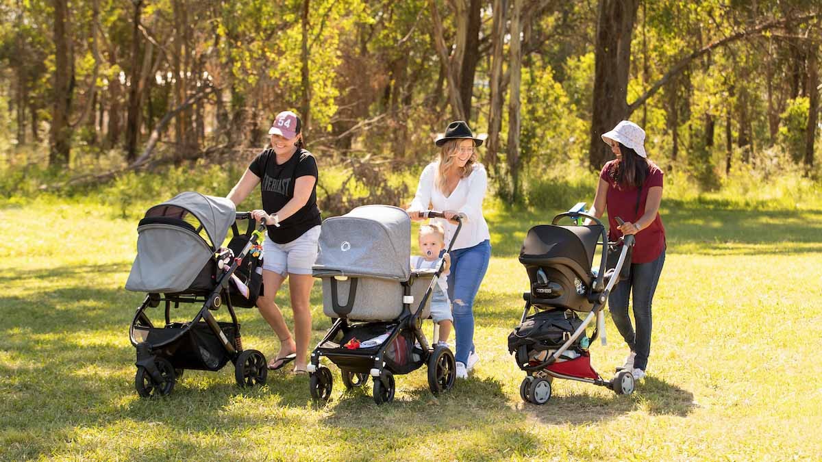 Three adults each push a pram across a grass area with trees in the background.
