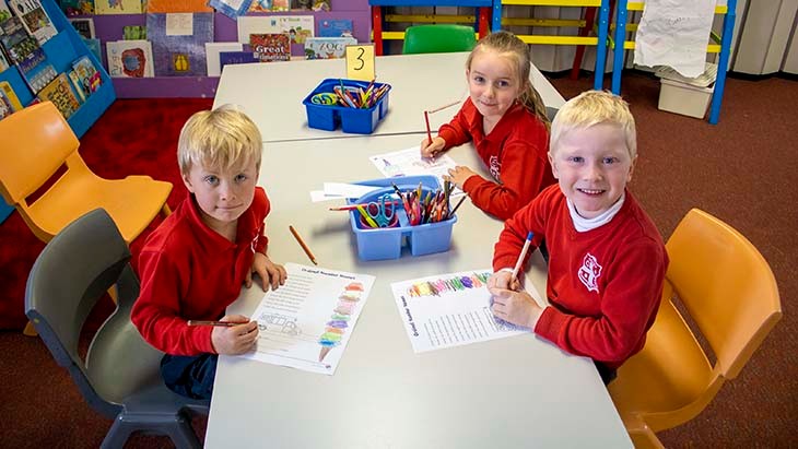 Three students wearing a red uniform sitting at a table colouring in.