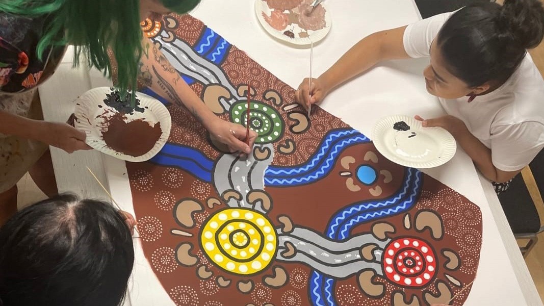 Three artists hard at work on a mural for the WorldPride Festival. The artwork is of an Aboriginal Design and features many dots, lines and circles in colours of blue, red, yellow and green.