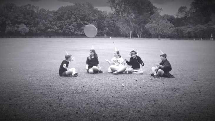 A black and white photo of five students sitting on the grass wearing party hats with a balloon floating above them.