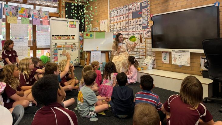 A teacher at the front of a classroom with students sitting on the floor.