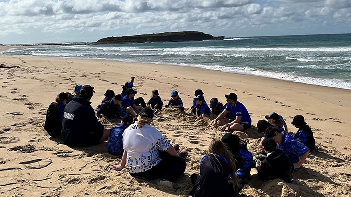 A group of children sit in a circle at the beach with an island in the background