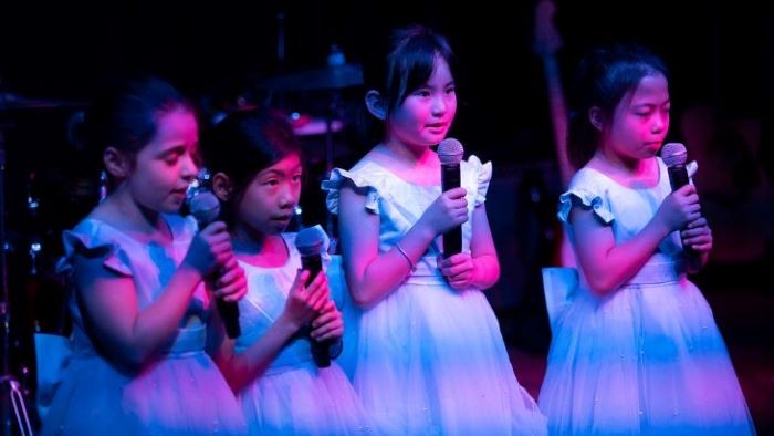 Four young children dressed in white dresses and singing into a microphone