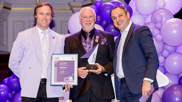 Three men posing for a photo in front of purple balloons. One man is holding a certificate, another a trophy.