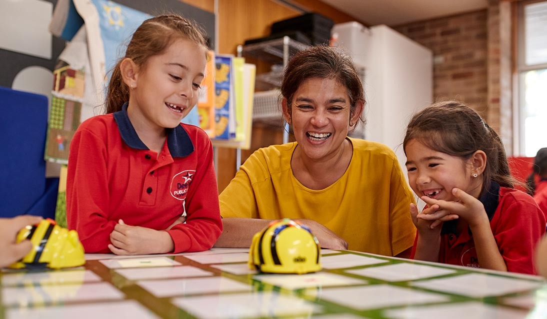 Two primary school girls and a teacher play with beebots in the classroom.