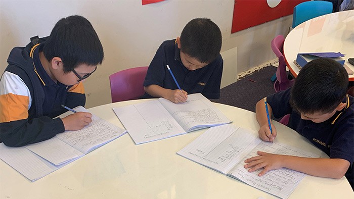 Three boys sitting at an oval table writing in their schoolbooks