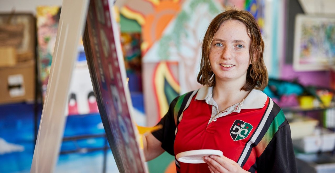 Female student painting at an easel. The background has many student colourful artworks on the wall. 