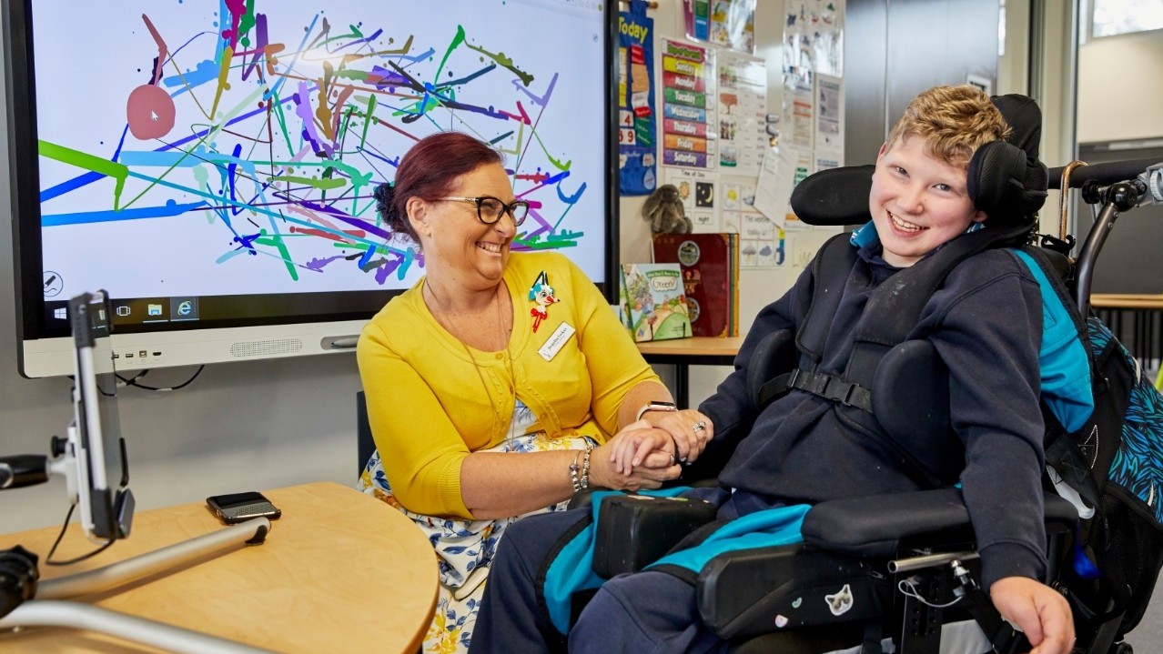 Teacher sitting next to student in a wheelchair in a classroom equipped with a range of specialist equipment.