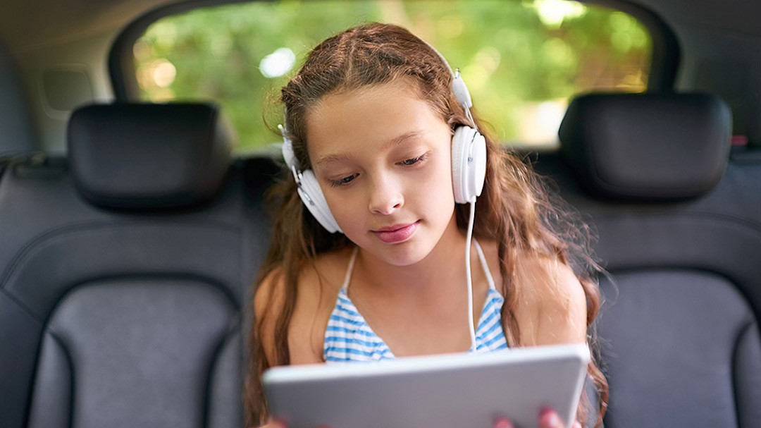 A young girl in a car, using headphones with her device.