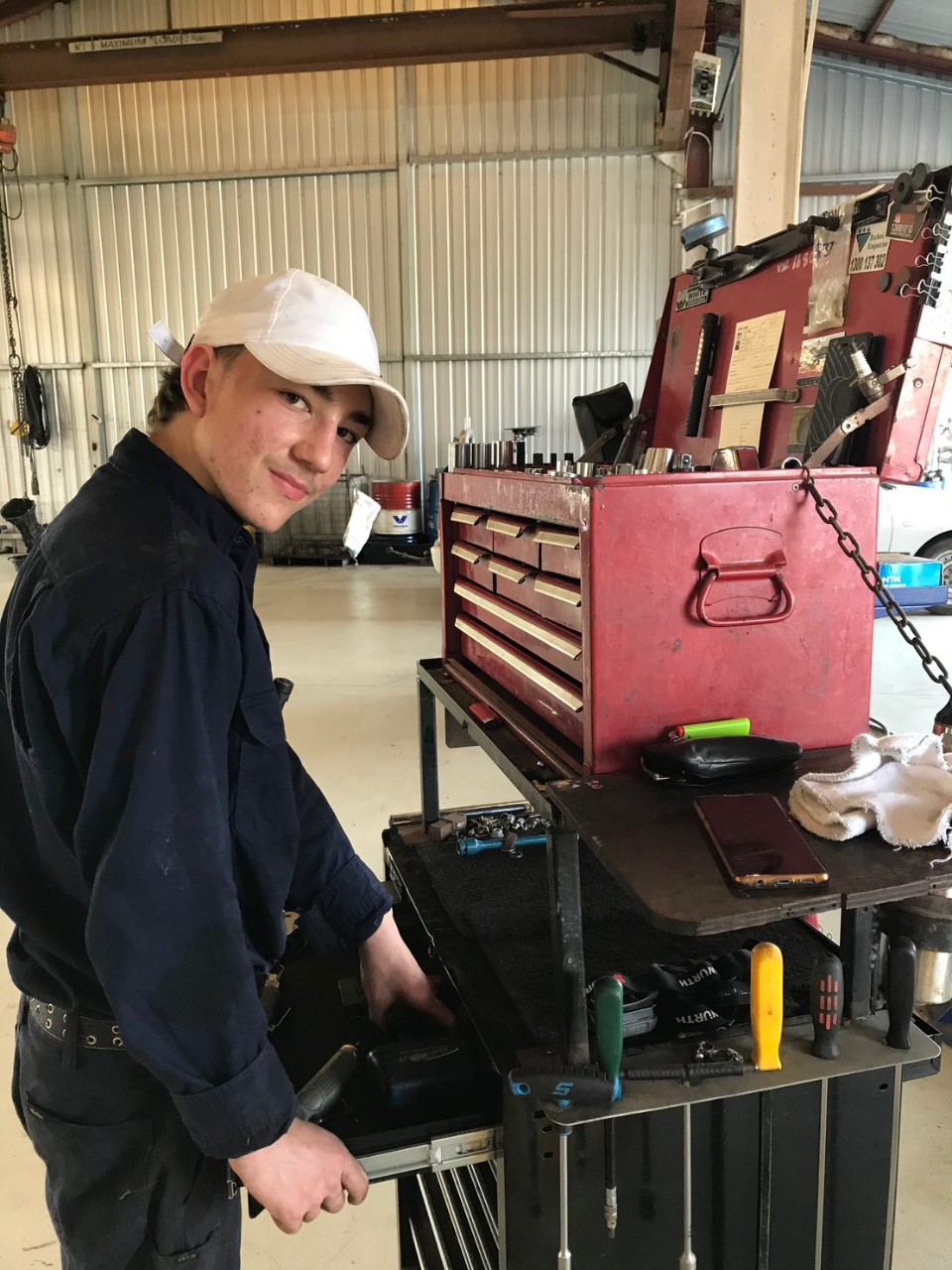 Photo of student with a white hat, and black collared shirt and black pants. He is smiling, and opening a red toolbox in a workshop