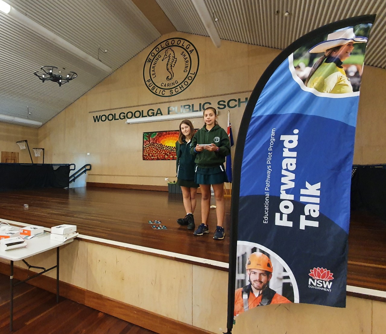 Two students standing on stage in school hall controlling flying a drone