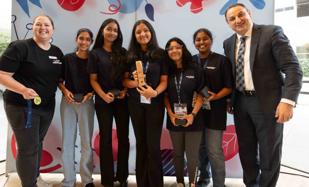Adult female stands in a line next to five female students. One student is holding a wooden trophy. An adult male stands on the end.