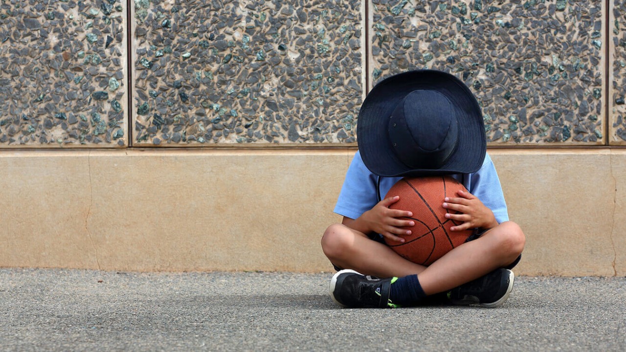 Child sitting on the ground, holding a basketball, with their face hidden by a hat