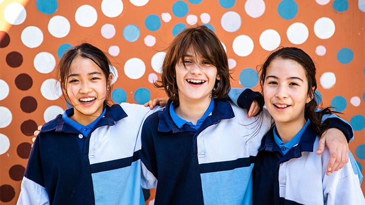 Three primary school students with arms around each other smiling in front of wall with Aboriginal art