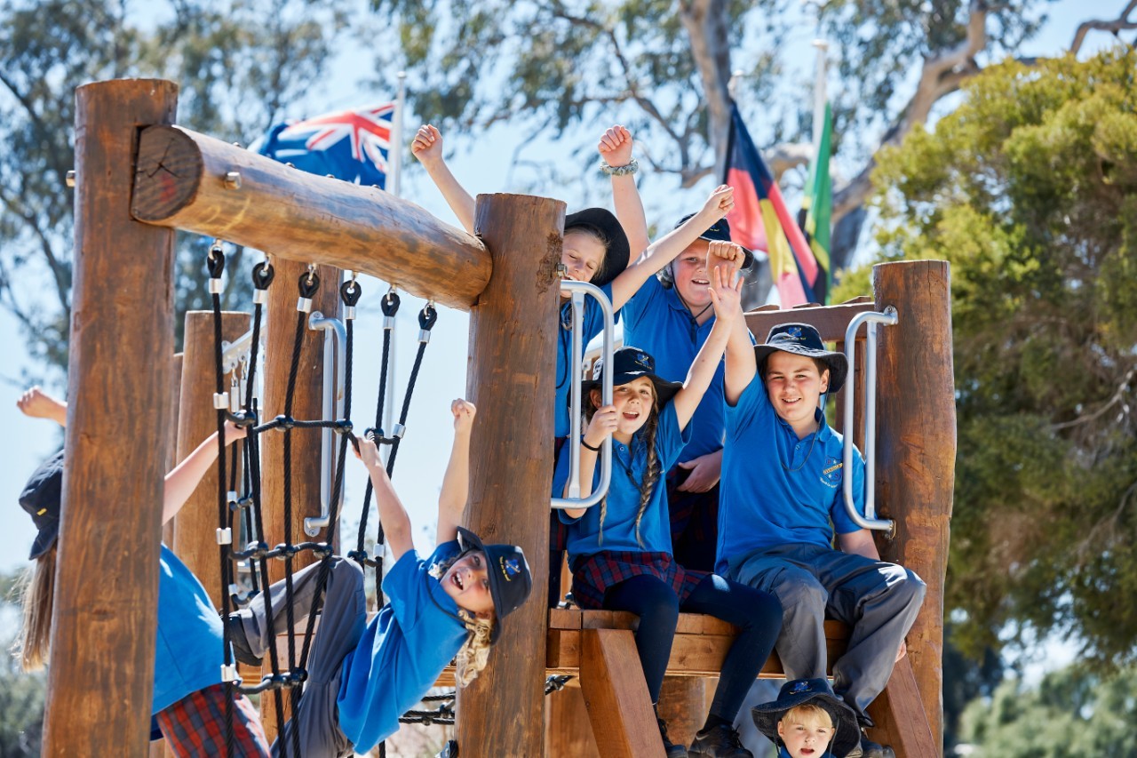 Group of young students wearing blue school uniform smile with their arms raised while they sit on outdoor play equipment.