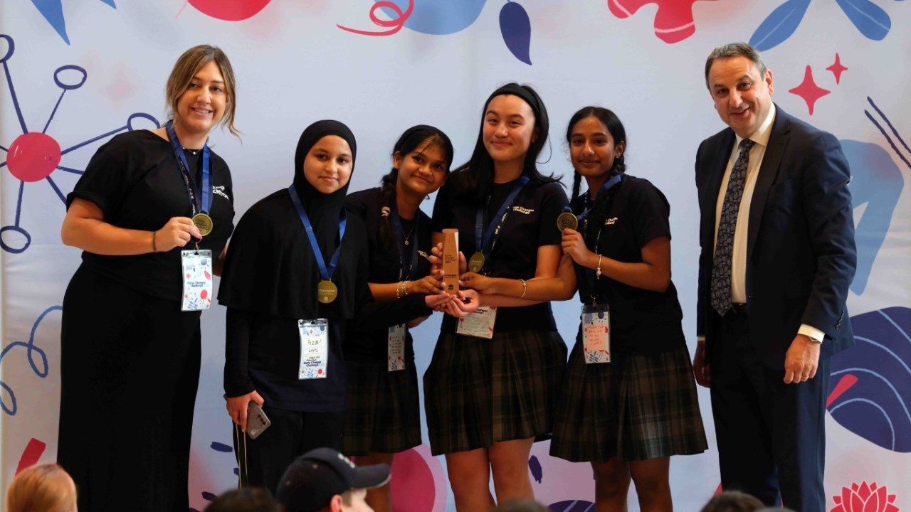 Four female students stand smiling in a line holding a wooden trophy. A male wearing a suit stands on the end and a female teacher stands at the other end.