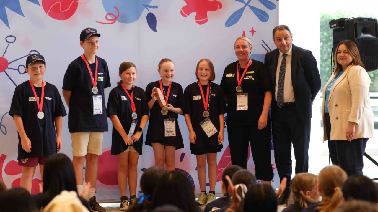 Five students stand smiling in a line. A male adult stands in between two female adults, next to the students. One student is holding a wooden trophy.