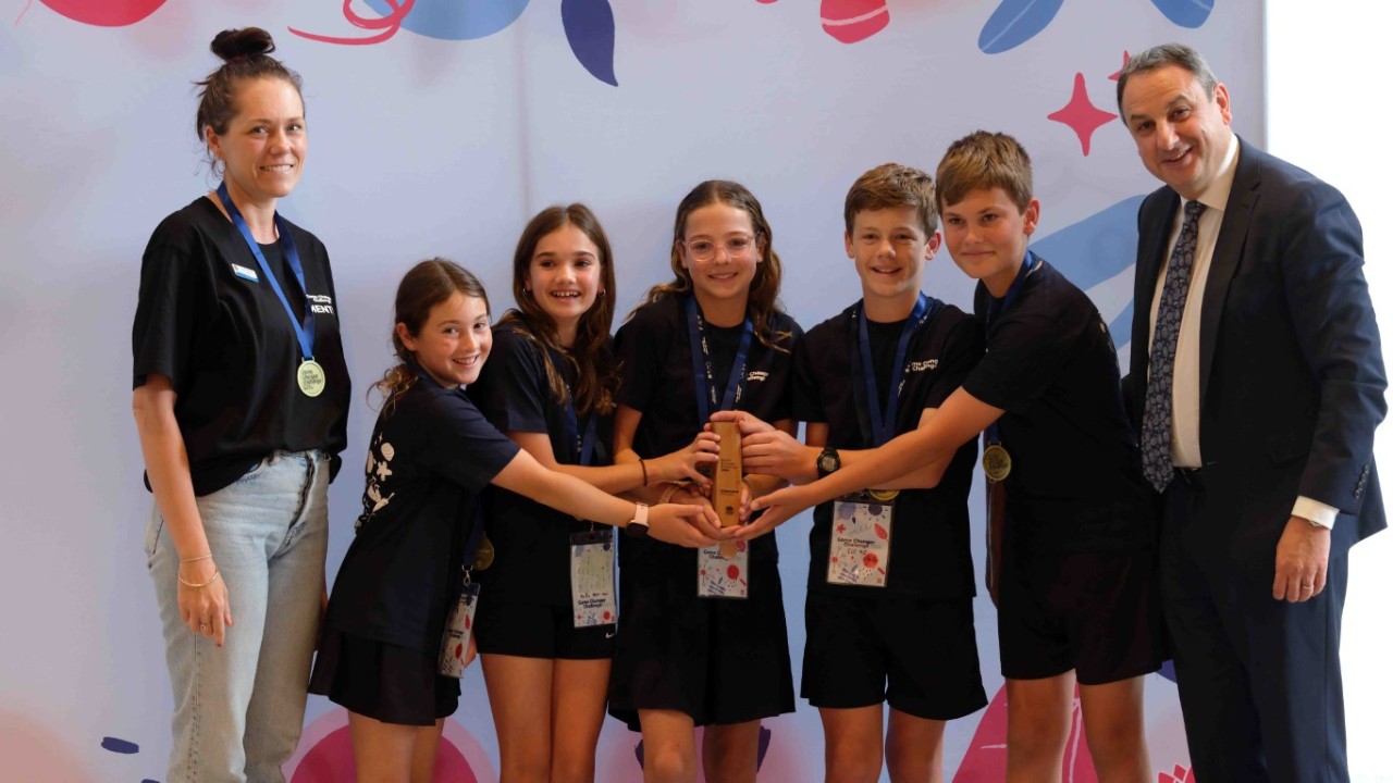 Five students stand in a line reaching arms out to hold a wooden trophy. A male and female adult stand on either side of the students.