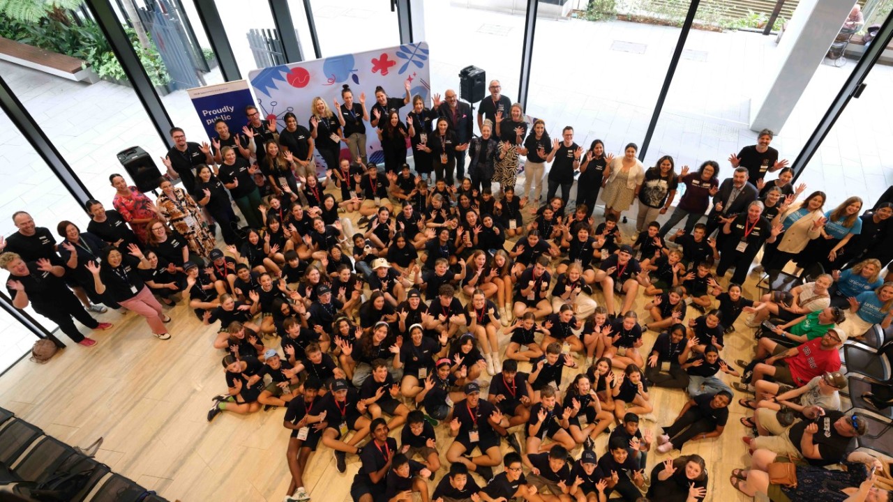 A large group of 100 people look up towards a camera and wave.