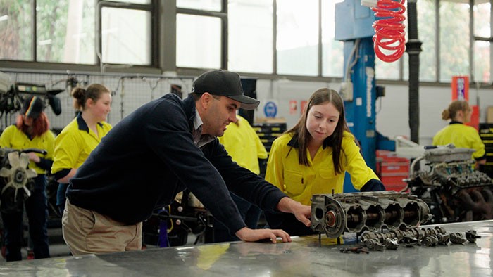 Female year 10 and 11 students being shown an engine in a mechanics workshop.