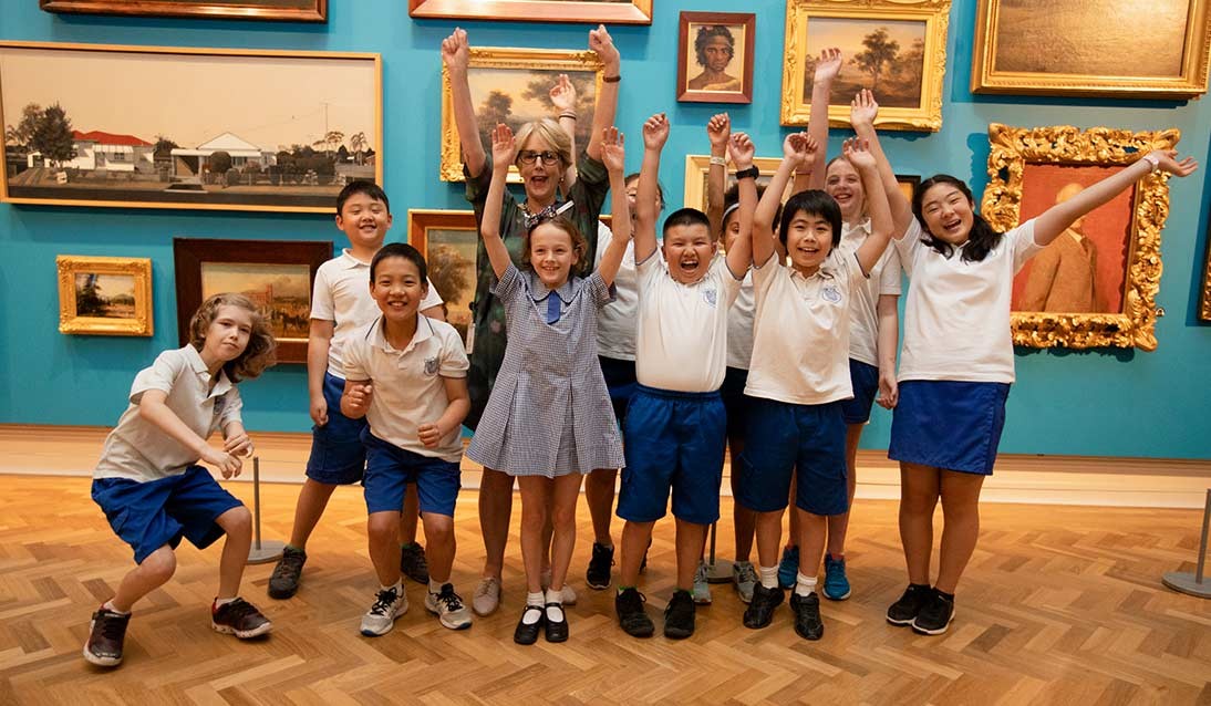 A group of primary-aged students and an adult raise their arms in excitement in front of a collection of artworks on a wall
