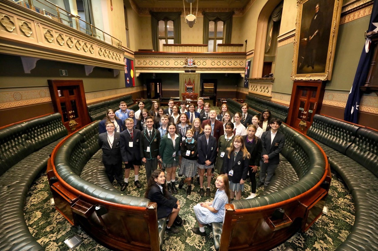 A group of students stand together in parliament house, with two students seated on benches
