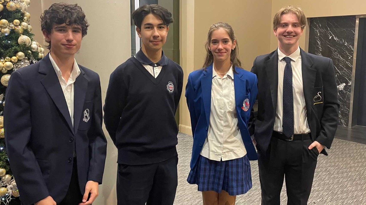 Four students smile as they stand in a line next to each other. Two male students, one female student and a third male. The students are wearing school uniform blazers in colours of black, navy and blue.