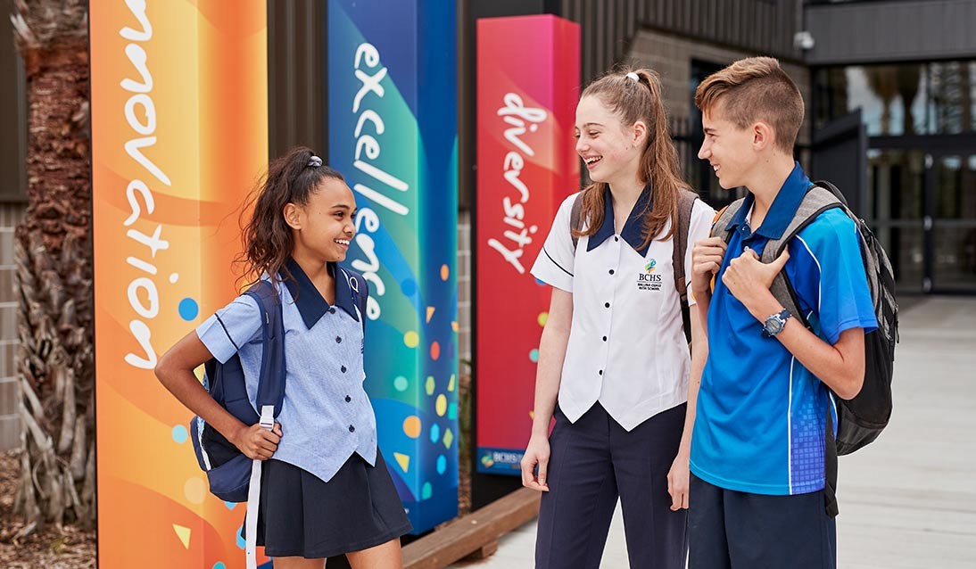 Three students in uniform stand outside talking to each other in front of three coloured pillars with the words innovation, excellence and diversity printed on them.