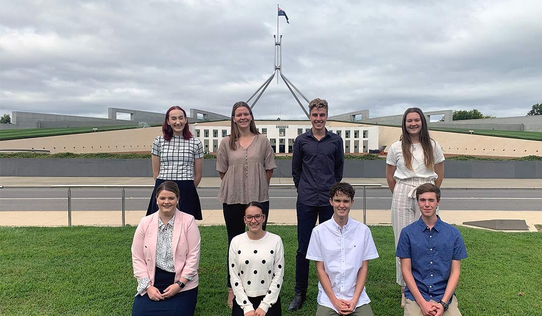 A group of eight people standing with the National Parliament of Australia building in the background.