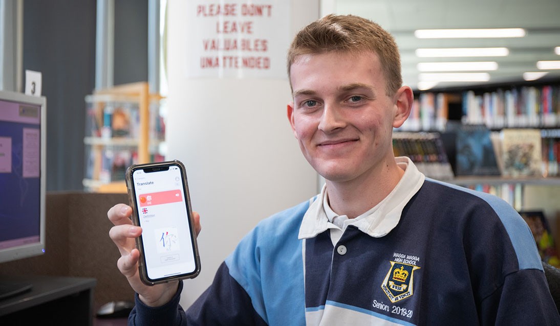 Male student wearing school uniform holds a mobile phone with his right hand. Phone screen shows language app interface.