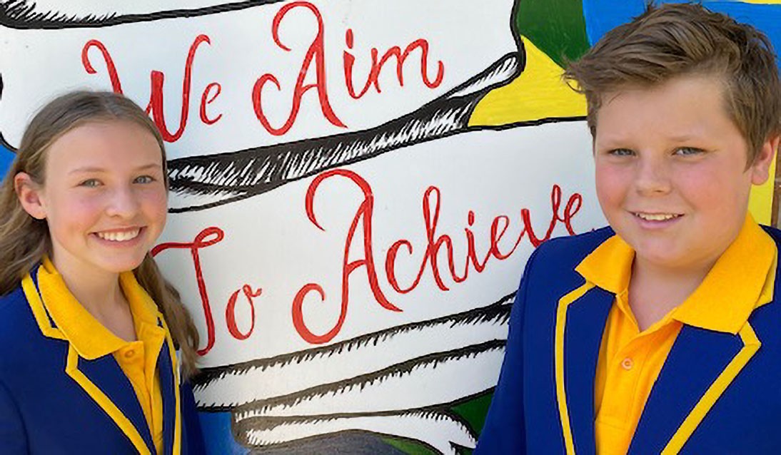 Female and male student wearing blue and yellow school uniform stand in front of mural with words We Aim to Achieve.
