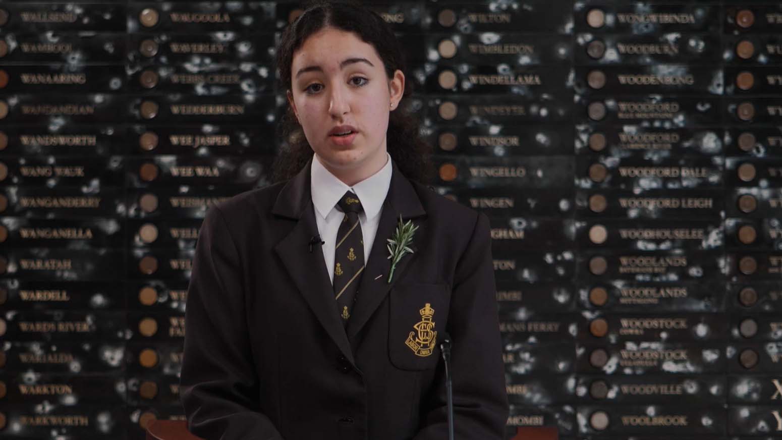 Female student wearing school blazer and rosemary sprig stands in front of a marble wall.