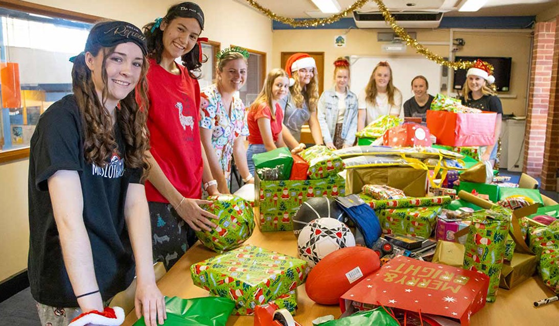Students stand around wrapped Christmas presents sitting on a table.