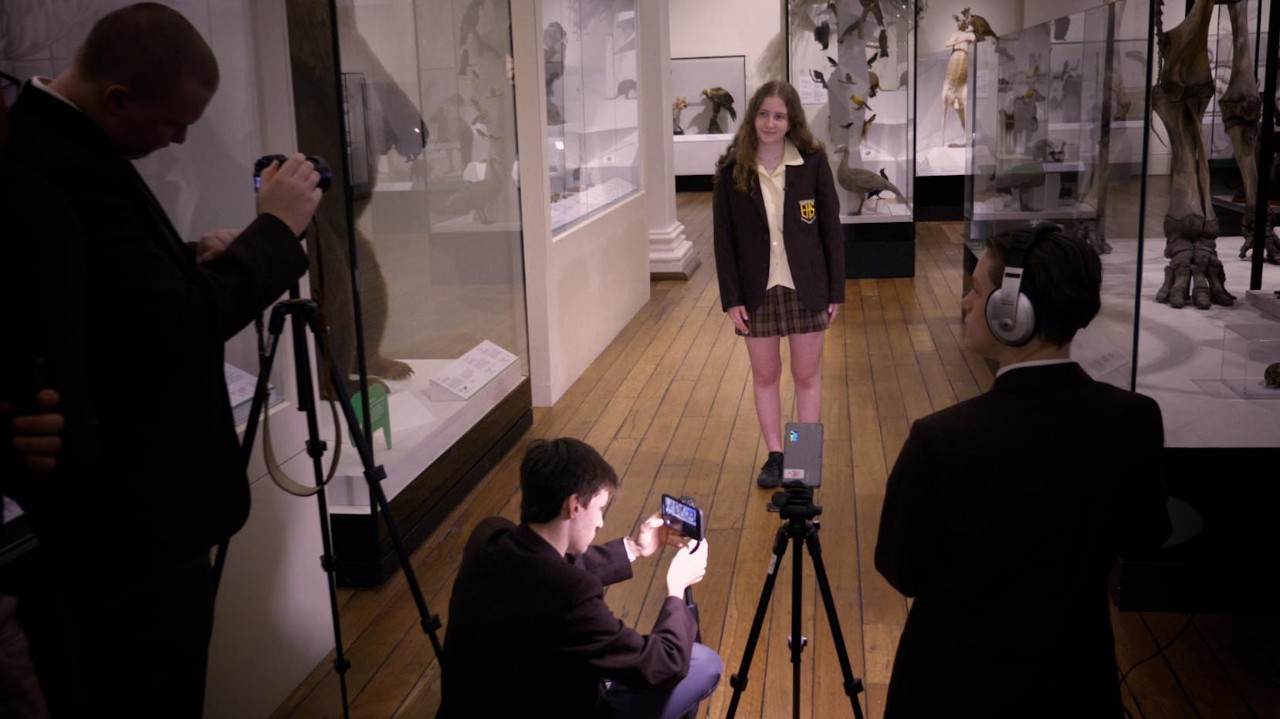 Student stands in room with museum exhibits in front of camera with students filming.