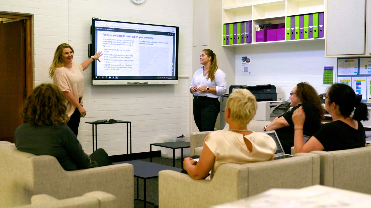 Two teachers are presenting from a digital board to four colleagues seated around the room in armchairs
