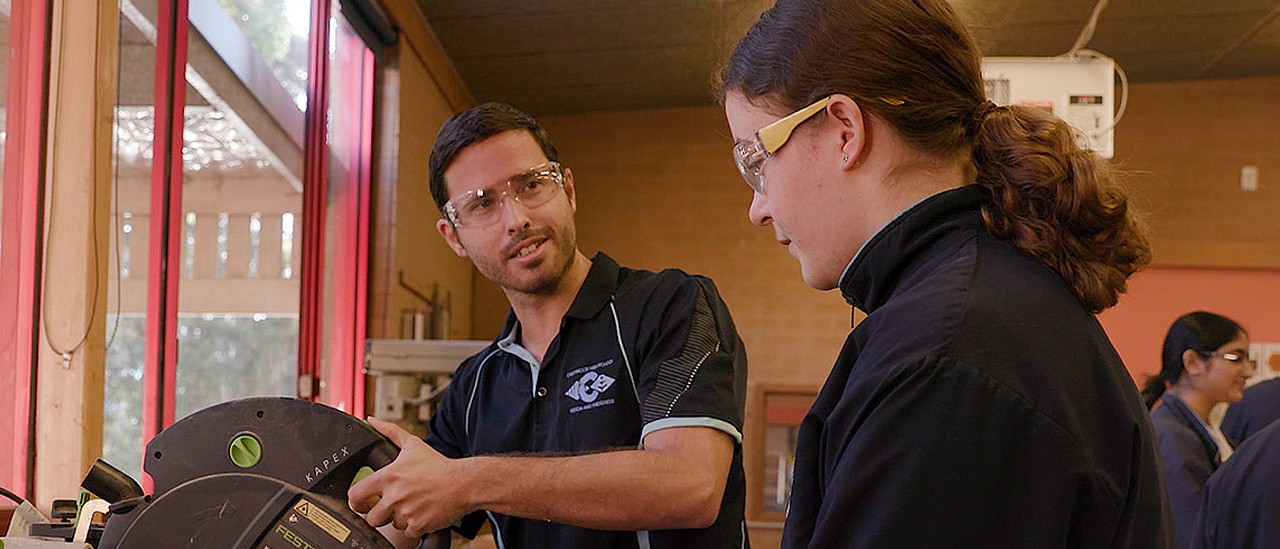 Male teacher in workshop demonstrating how to use machinery with female student