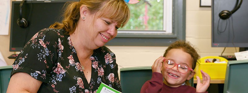 Female teacher sits to the left of a smiling student reading a book