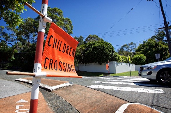 Image of an orange Children Crossing sign in the foreground with a zebra crossing and car in the background