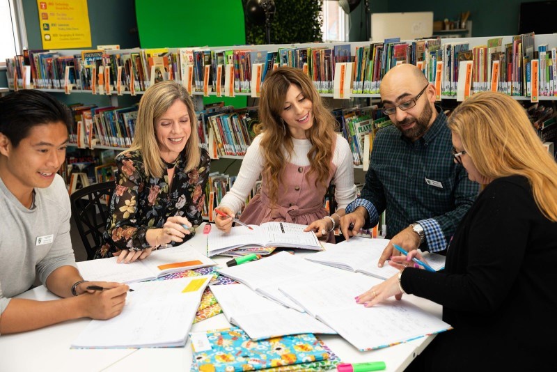 A group of five teachers comprising beginning teachers and their supervisors or in school mentors are engaged in a round table discussion together in the school library  They have a number of samples of students work open in front of them which they are reviewing and analysing.