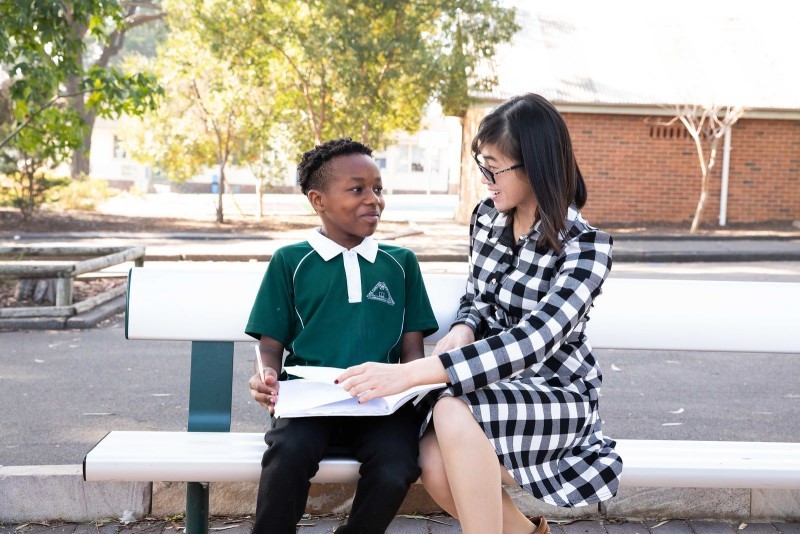 A beginning teacher is sitting next to a male student in a school playground setting. He is holding his exercise book open on his lap whilst she is providing him with oral feedback on his work.