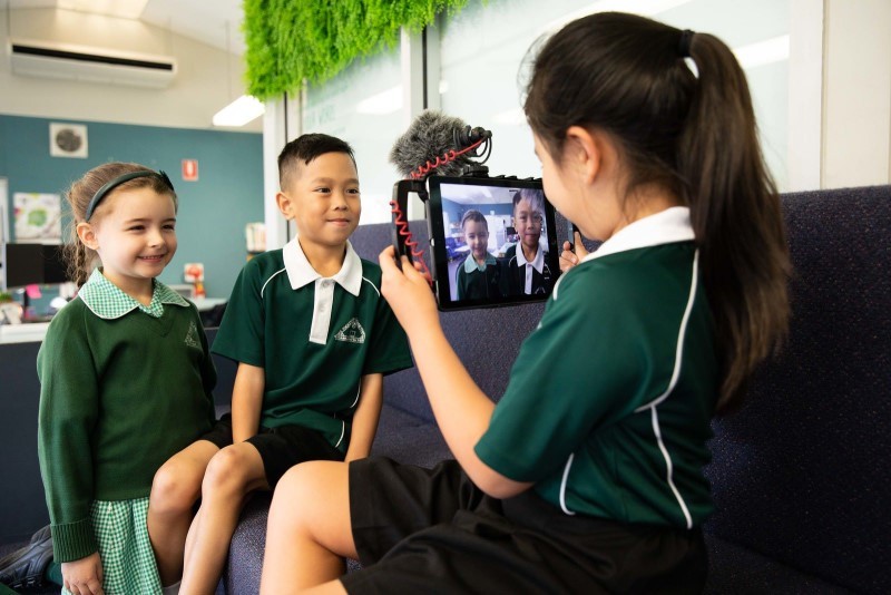 Three primary students, two girls and one boy are participating in a lesson. One of the girls is filming the other two students as they complete the tasks for the lesson.