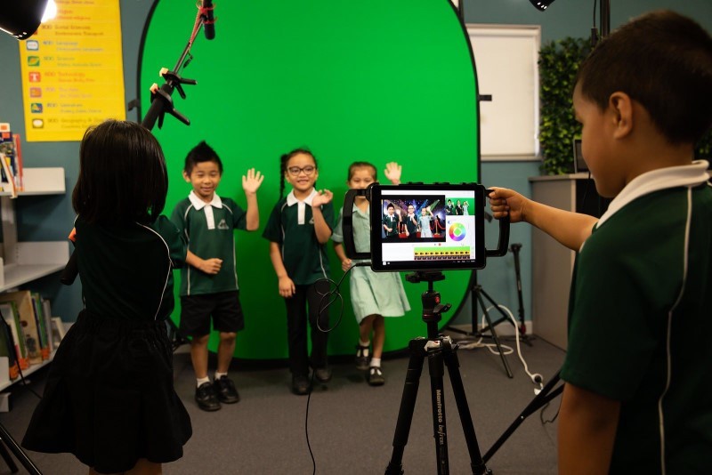 Group of five junior students in front of a plain green screen making a movie in a classroom setting. One male student is in front of a camera on a tripod. The camera is focused on three students who are waving directly at the camera. A fifth student is in the wings holding a microphone on a boom arm up to the three students being filmed.