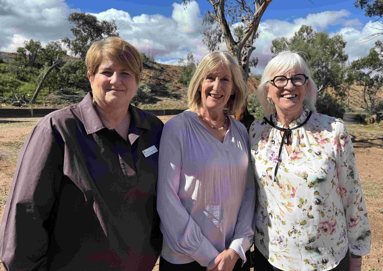 Three women are standing in the outback in front of some trees.
