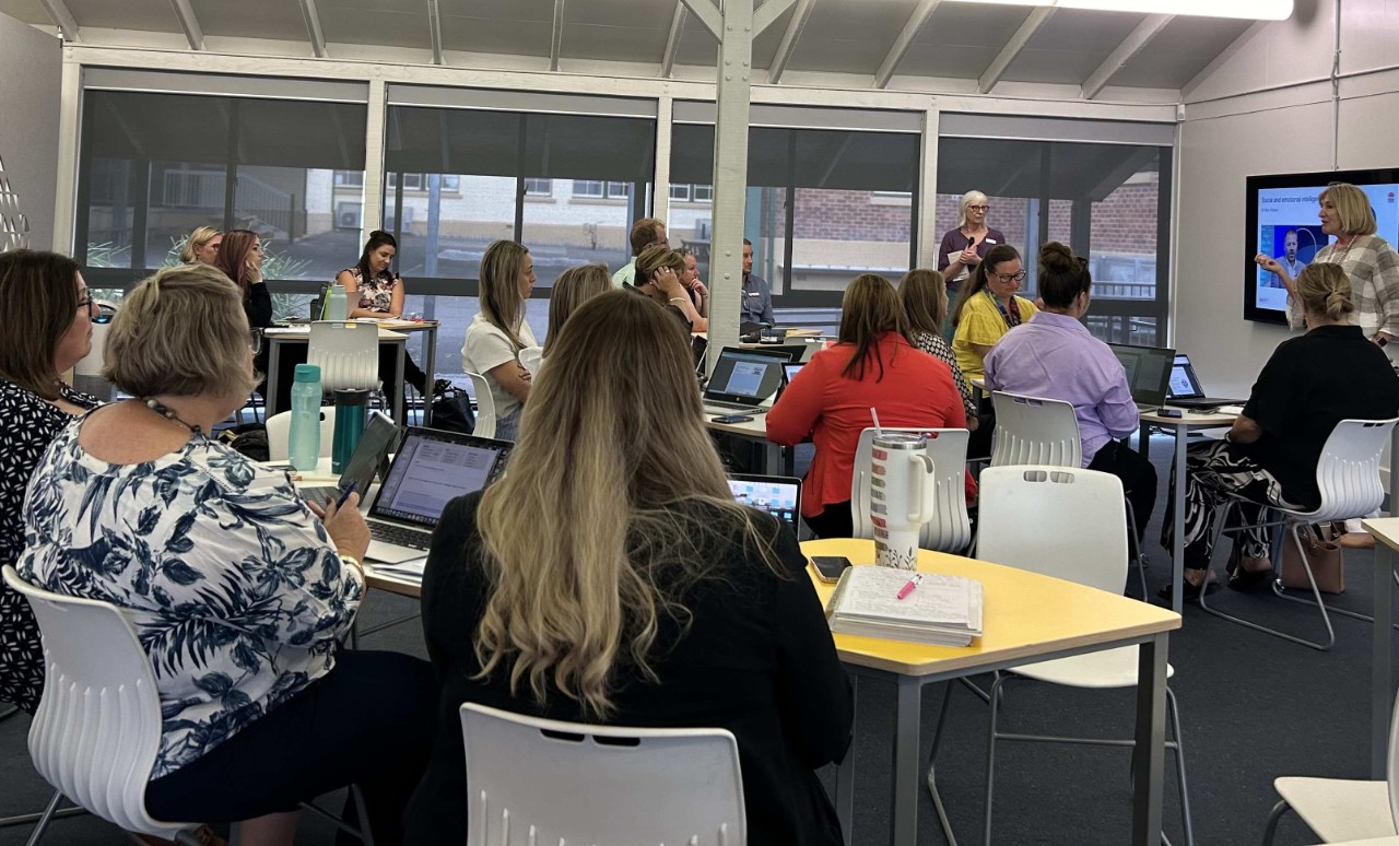 A group of educators sit in a classroom with presenters at the front of the room delivering professional learning.